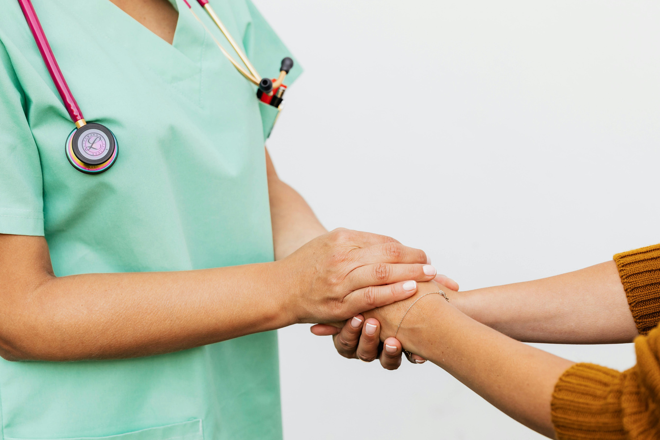 A nurse in teal scrubs with a stethoscope gently holds a patient's hand during a recuperative-care check-in